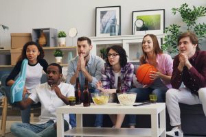 A group of young friends enthusiastically watching a sports event indoors with snacks and drinks.