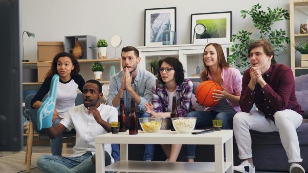 A group of young friends enthusiastically watching a sports event indoors with snacks and drinks.