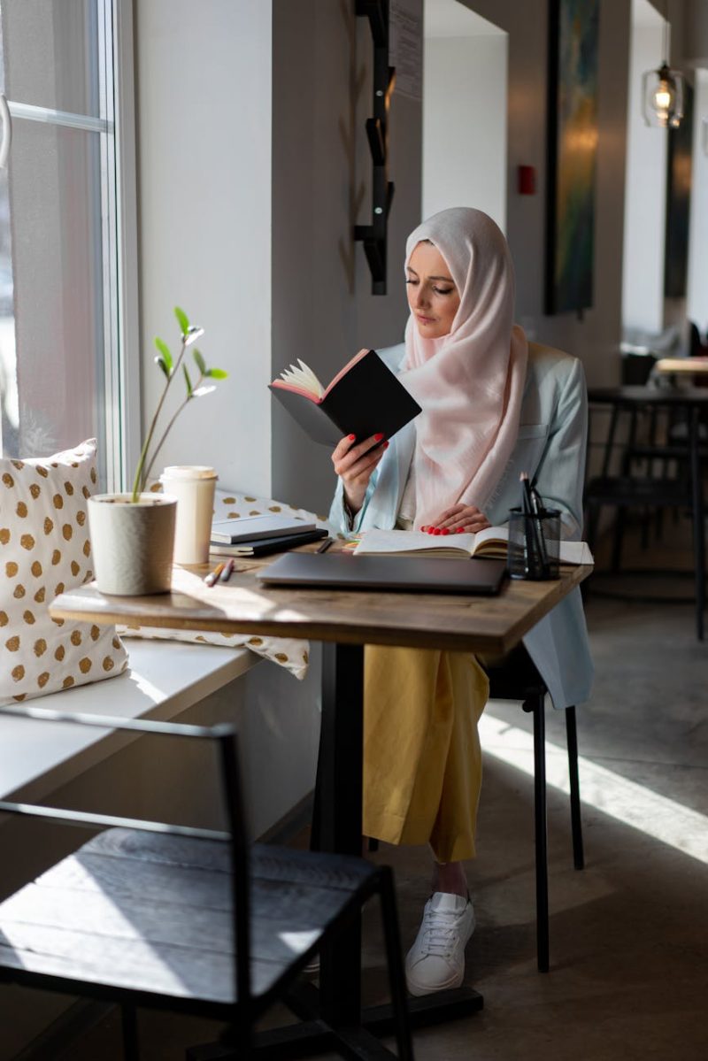 A young woman in a hijab reading a book at a cozy cafe table by the window.