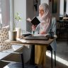 A young woman in a hijab reading a book at a cozy cafe table by the window.