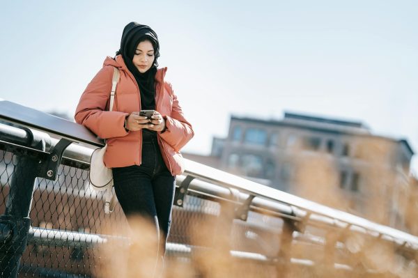 Low angle of young Muslim woman in casual outfit and traditional headscarf browsing mobile phone on street