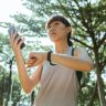 Young woman in sportswear checks smartwatch while using smartphone outdoors in a sunny park.