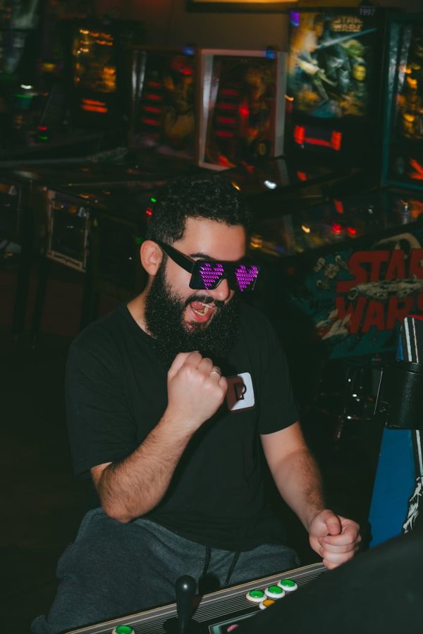 A man celebrates victory in an arcade, wearing fun glasses and a beard.