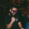 A man celebrates victory in an arcade, wearing fun glasses and a beard.