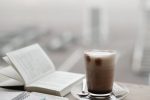 A warm coffee sits beside a notebook and smartphone on a foggy day.