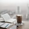 A warm coffee sits beside a notebook and smartphone on a foggy day.
