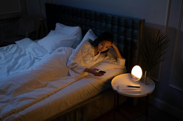 An Asian woman on a bed using her phone in a dim bedroom, reflecting on insomnia or night routines.